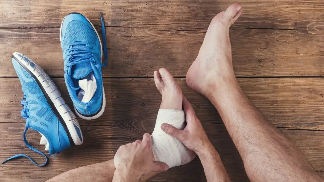 Skręcona kostka z opuchlizną, owinięta bandażem. Obok leżą niebieskie buty sportowe.
