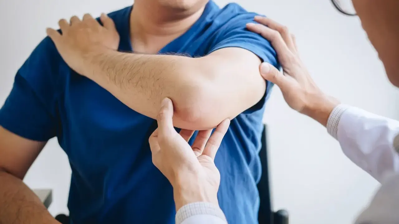 Fizjoterapeuta bada łokieć pacjenta. Dłonie specjalisty delikatnie dotykają bolącego miejsca, oceniając zakres ruchu i ból.