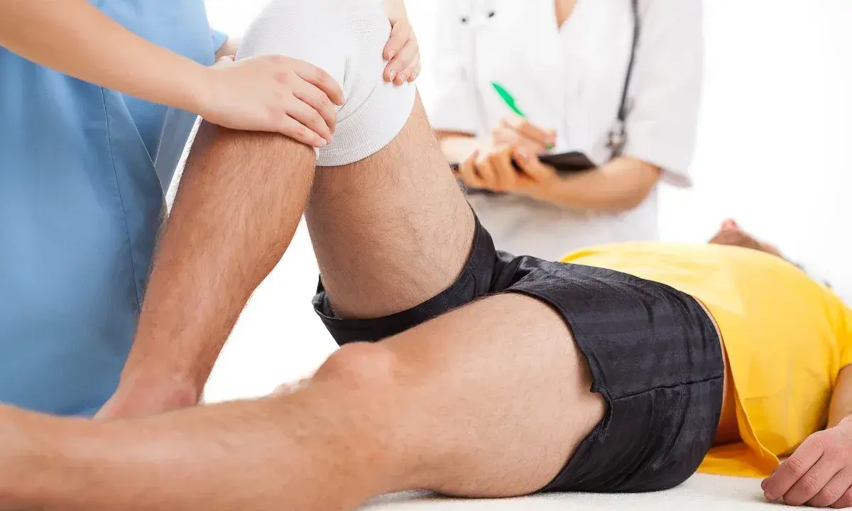 Fizjoterapia kolana na czym polega? Terapeuta delikatnie uciska opatrunek na kolanie pacjenta, który leży na stole.