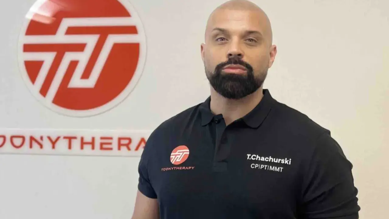 Tomasz Chachurski w czarnej koszulce z logo Toonytherapy, na tle białej ściany z logo.