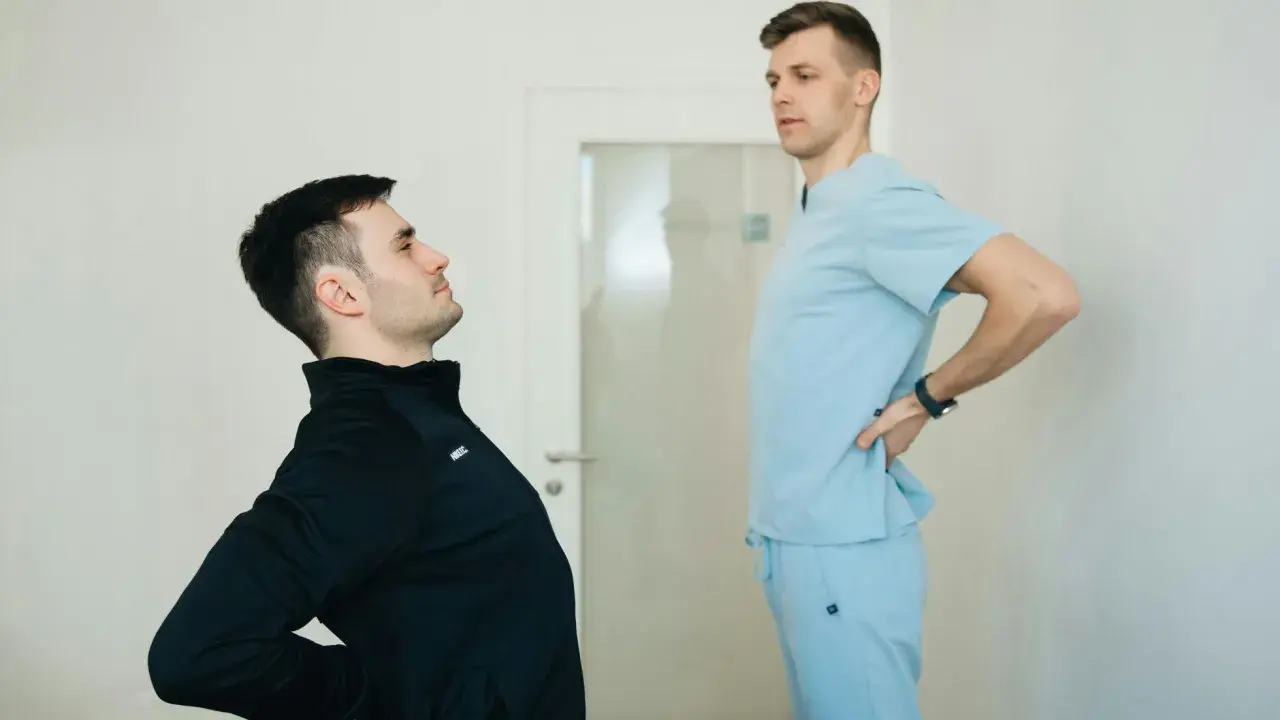 Fizjoterapeuta w jasnoniebieskim uniformie rozmawia z pacjentem, który odczuwa ból pleców. Dowiedz się, ile kosztuje wizyta fizjoterapeuty w domu.