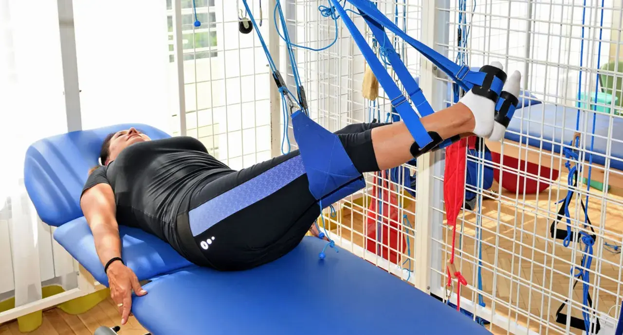 Kobieta na łóżku rehabilitacyjnym ćwiczy nogi w systemie podwieszeń. To przykład, jak wygląda rehabilitacja dzienna co to jest.