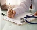 Lekarz pisze receptę, dokumentując choroby, na jakie można dostać orzeczenie o niepełnosprawności. Stetoskop i laptop w tle.