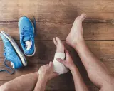 Skręcona kostka z opuchlizną, owinięta bandażem. Obok leżą niebieskie buty sportowe.