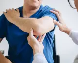 Fizjoterapeuta bada łokieć pacjenta. Dłonie specjalisty delikatnie dotykają bolącego miejsca, oceniając zakres ruchu i ból.