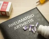 Na czarnej okładce z napisem "RHEUMATOID ARTHRITIS" leżą kapsułki. Obok znajduje się pudełko leków i strzykawka, sugerujące najnowsze leki na reumatoidalne zapalenie stawów.