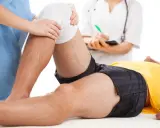 Fizjoterapia kolana na czym polega? Terapeuta delikatnie uciska opatrunek na kolanie pacjenta, który leży na stole.