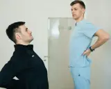 Fizjoterapeuta w jasnoniebieskim uniformie rozmawia z pacjentem, który odczuwa ból pleców. Dowiedz się, ile kosztuje wizyta fizjoterapeuty w domu.