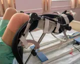 Pacjent po zabiegu z endoprotezą kolana w specjalistycznym urządzeniu rehabilitacyjnym, z bandażem na kolanie.