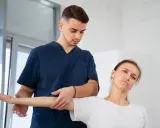 Młody fizjoterapeuta wykonuje badanie ręki pacjentki. Czy można zostać fizjoterapeutą bez studiów?