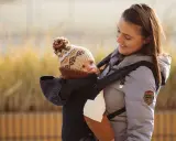 Fizjoterapeuta poleca nosidełko dla niemowlaka, które zapewnia prawidłową pozycję bioder i kręgosłupa. Uśmiechnięta mama z dzieckiem w nosidełku.
