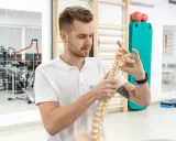 Fizjoterapeuta pokazuje model kręgosłupa, wyjaśniając, czy fizjoterapia jest trudna. W tle sprzęt rehabilitacyjny.