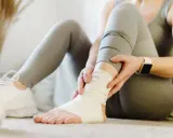 Kobieta owija bandażem skręconą kostkę, siedząc na podłodze w sportowych legginsach i butach.