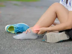 Ból kostki po bieganiu, czerwony obszar wskazuje na zwichnięcie. Obok leżą niebieskie buty sportowe.