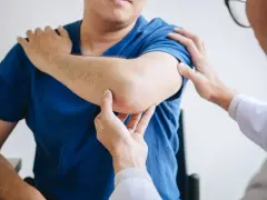 Fizjoterapeuta bada łokieć pacjenta. Dłonie specjalisty delikatnie dotykają bolącego miejsca, oceniając zakres ruchu i ból.