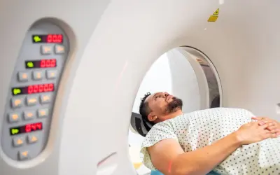 Pacjent leży w aparacie do tomografii komputerowej, przygotowując się do badania. Obok widoczny panel sterowania z cyfrowymi wskaźnikami.