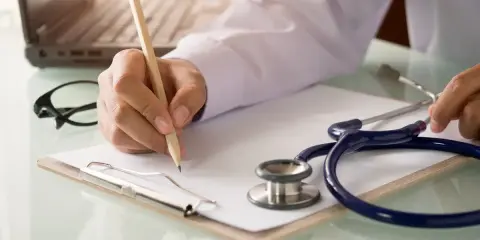 Lekarz pisze receptę, dokumentując choroby, na jakie można dostać orzeczenie o niepełnosprawności. Stetoskop i laptop w tle.