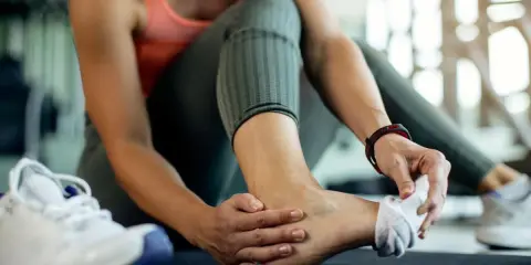 Kobieta masuje bolącą kostkę w siłowni, obok leży but sportowy.