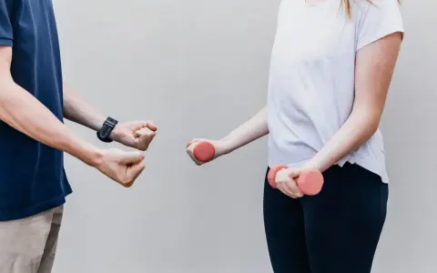 Dwie osoby ćwiczą z hantlami. Jedna osoba pokazuje zaciśniętą pięść, druga trzyma różowe hantle. Z czym do fizjoterapeuty?