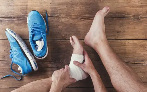 Skręcona kostka z opuchlizną, owinięta bandażem. Obok leżą niebieskie buty sportowe.