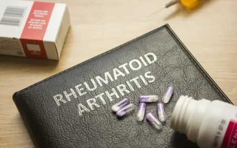 Na czarnej okładce z napisem "RHEUMATOID ARTHRITIS" leżą kapsułki. Obok znajduje się pudełko leków i strzykawka, sugerujące najnowsze leki na reumatoidalne zapalenie stawów.