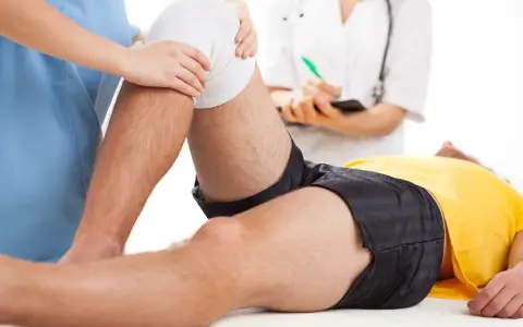 Fizjoterapia kolana na czym polega? Terapeuta delikatnie uciska opatrunek na kolanie pacjenta, który leży na stole.