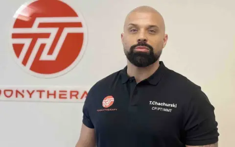 Tomasz Chachurski w czarnej koszulce z logo Toonytherapy, na tle białej ściany z logo.