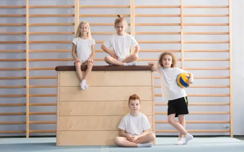 Dzieci w sali gimnastycznej. Czy fizjoterapeuta może wystawić zwolnienie z wf? Dwoje siedzi na skrzyni, jedno na niej, jedno na podłodze.
