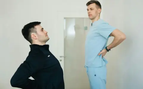 Fizjoterapeuta w jasnoniebieskim uniformie rozmawia z pacjentem, który odczuwa ból pleców. Dowiedz się, ile kosztuje wizyta fizjoterapeuty w domu.