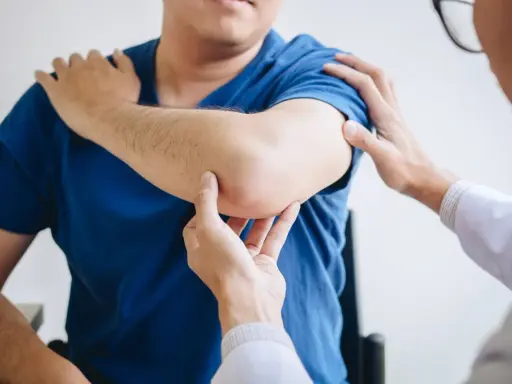 Fizjoterapeuta bada łokieć pacjenta. Dłonie specjalisty delikatnie dotykają bolącego miejsca, oceniając zakres ruchu i ból.