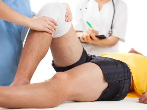 Fizjoterapia kolana na czym polega? Terapeuta delikatnie uciska opatrunek na kolanie pacjenta, który leży na stole.