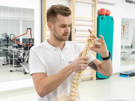 Fizjoterapeuta pokazuje model kręgosłupa, wyjaśniając, czy fizjoterapia jest trudna. W tle sprzęt rehabilitacyjny.