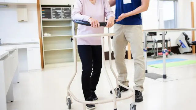 Osoba z niepełnosprawnością ćwiczy chodzenie z pomocą opiekuna i balkonika w sali rehabilitacyjnej.