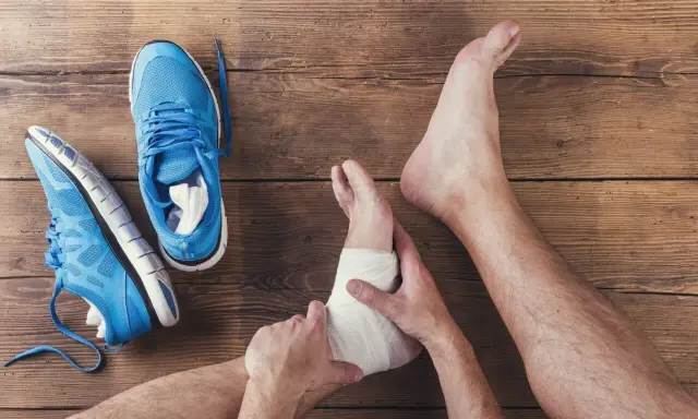 Skręcona kostka z opuchlizną, owinięta bandażem. Obok leżą niebieskie buty sportowe.