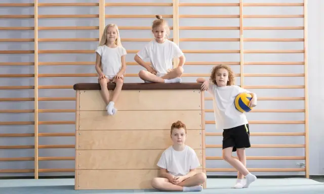 Dzieci w sali gimnastycznej. Czy fizjoterapeuta może wystawić zwolnienie z wf? Dwoje siedzi na skrzyni, jedno na niej, jedno na podłodze.