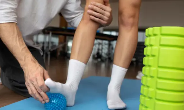 Fizjoterapeuta pomaga pacjentowi z niebieską piłeczką, co na skręconą kostkę. Obok zielony wałek do masażu.