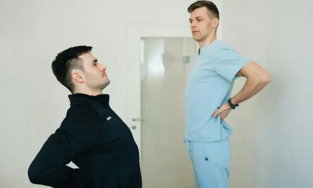 Fizjoterapeuta w jasnoniebieskim uniformie rozmawia z pacjentem, który odczuwa ból pleców. Dowiedz się, ile kosztuje wizyta fizjoterapeuty w domu.