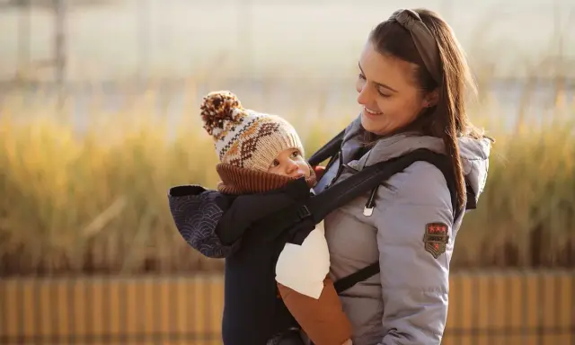 Fizjoterapeuta poleca nosidełko dla niemowlaka, które zapewnia prawidłową pozycję bioder i kręgosłupa. Uśmiechnięta mama z dzieckiem w nosidełku.