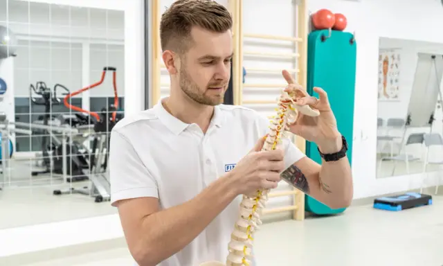 Fizjoterapeuta pokazuje model kręgosłupa, wyjaśniając, czy fizjoterapia jest trudna. W tle sprzęt rehabilitacyjny.