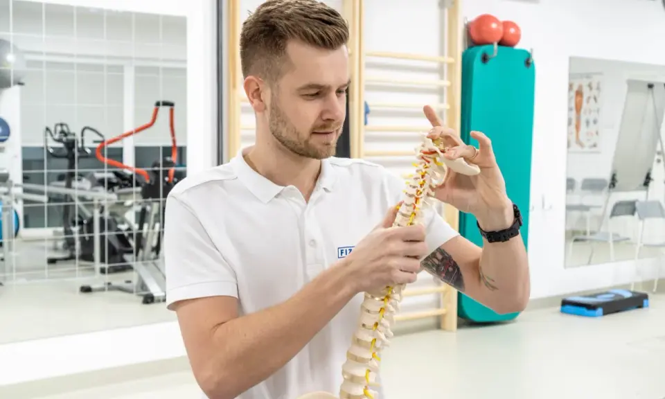 Fizjoterapeuta pokazuje model kręgosłupa, wyjaśniając, czy fizjoterapia jest trudna. W tle sprzęt rehabilitacyjny.