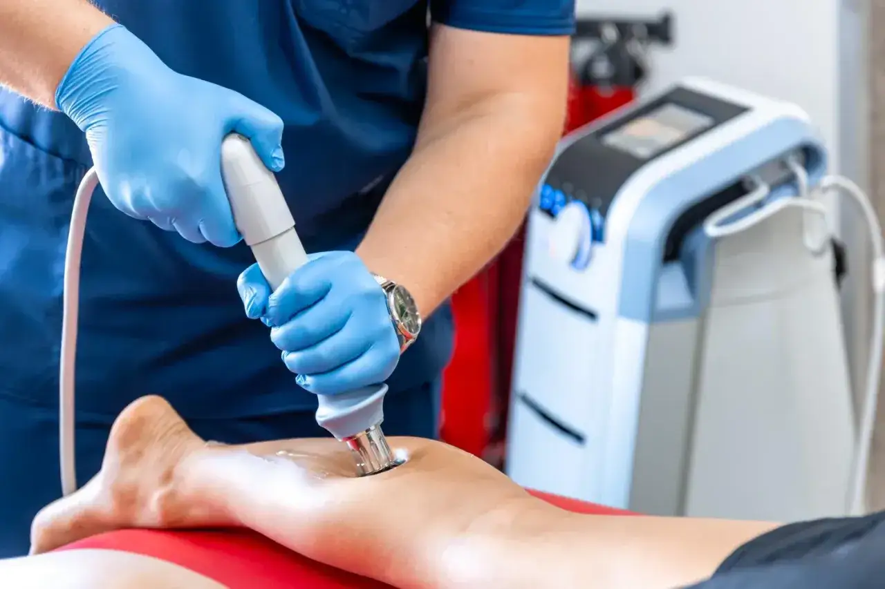 Fizjoterapeuta w rękawiczkach wykonuje rehabilitację laserem na nodze pacjenta. Zabieg ten wspomaga regenerację tkanek.