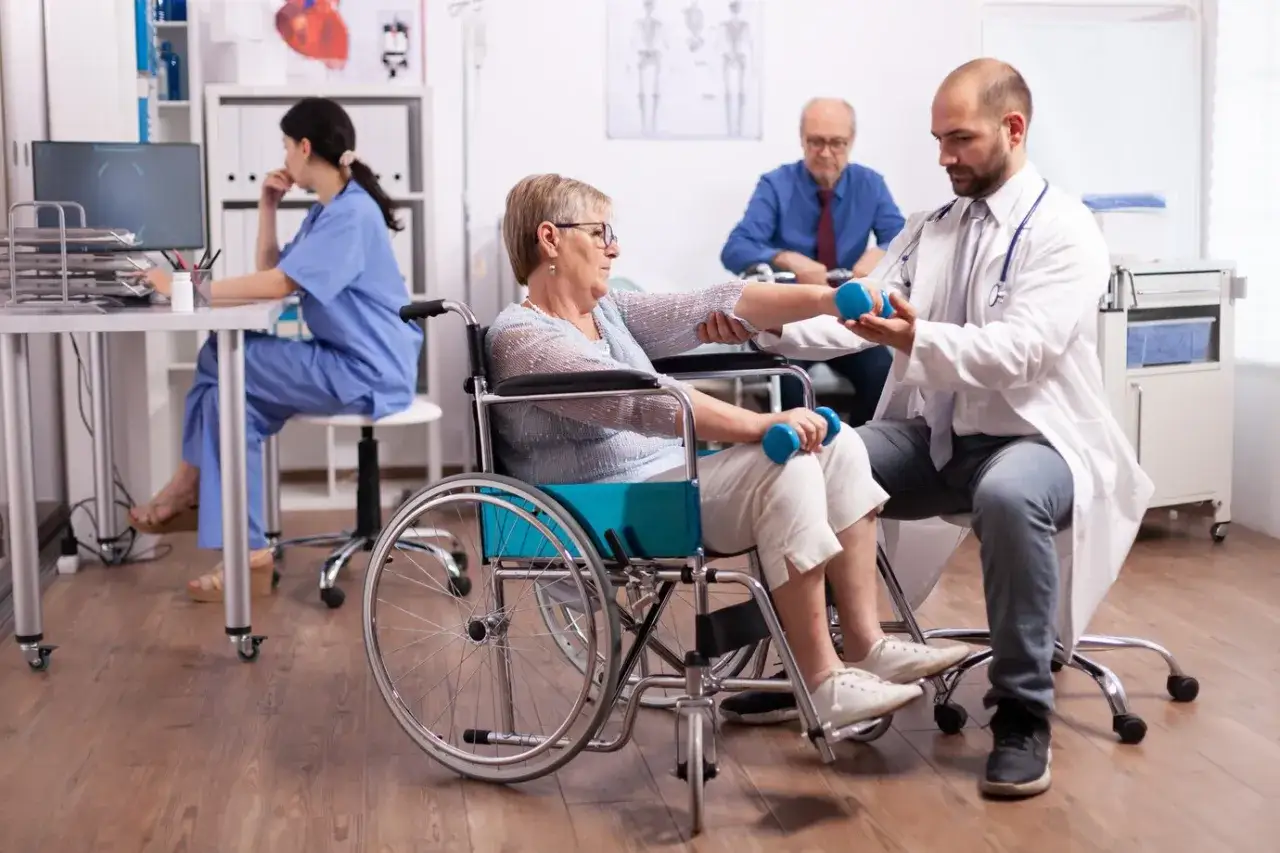 Rehabilitacja po udarze w szpitalu: pacjentka w wózku wykonuje ćwiczenia z hantlami pod okiem fizjoterapeuty.