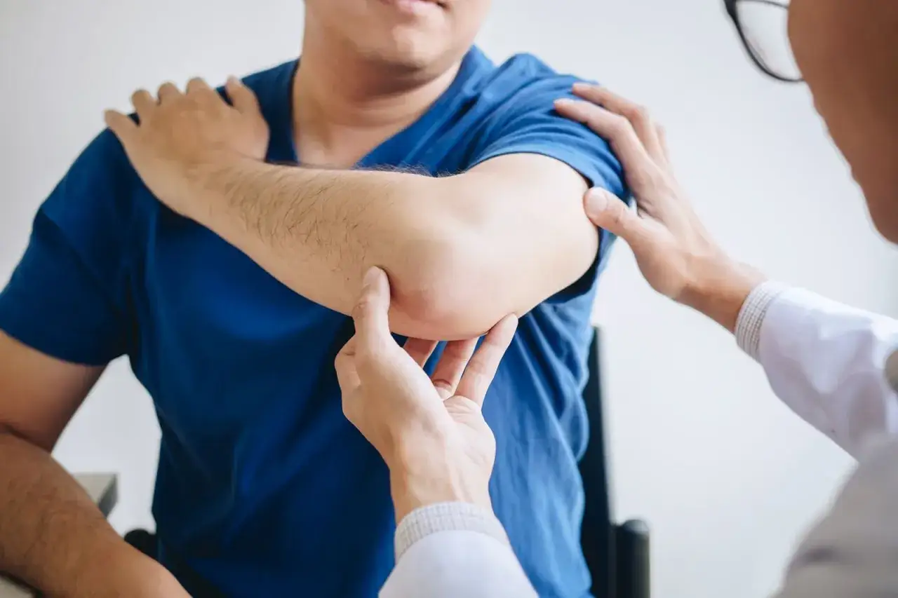 Fizjoterapeuta bada łokieć pacjenta. Dłoń specjalisty delikatnie uciska sk&oacute;rę, oceniając stan stawu.