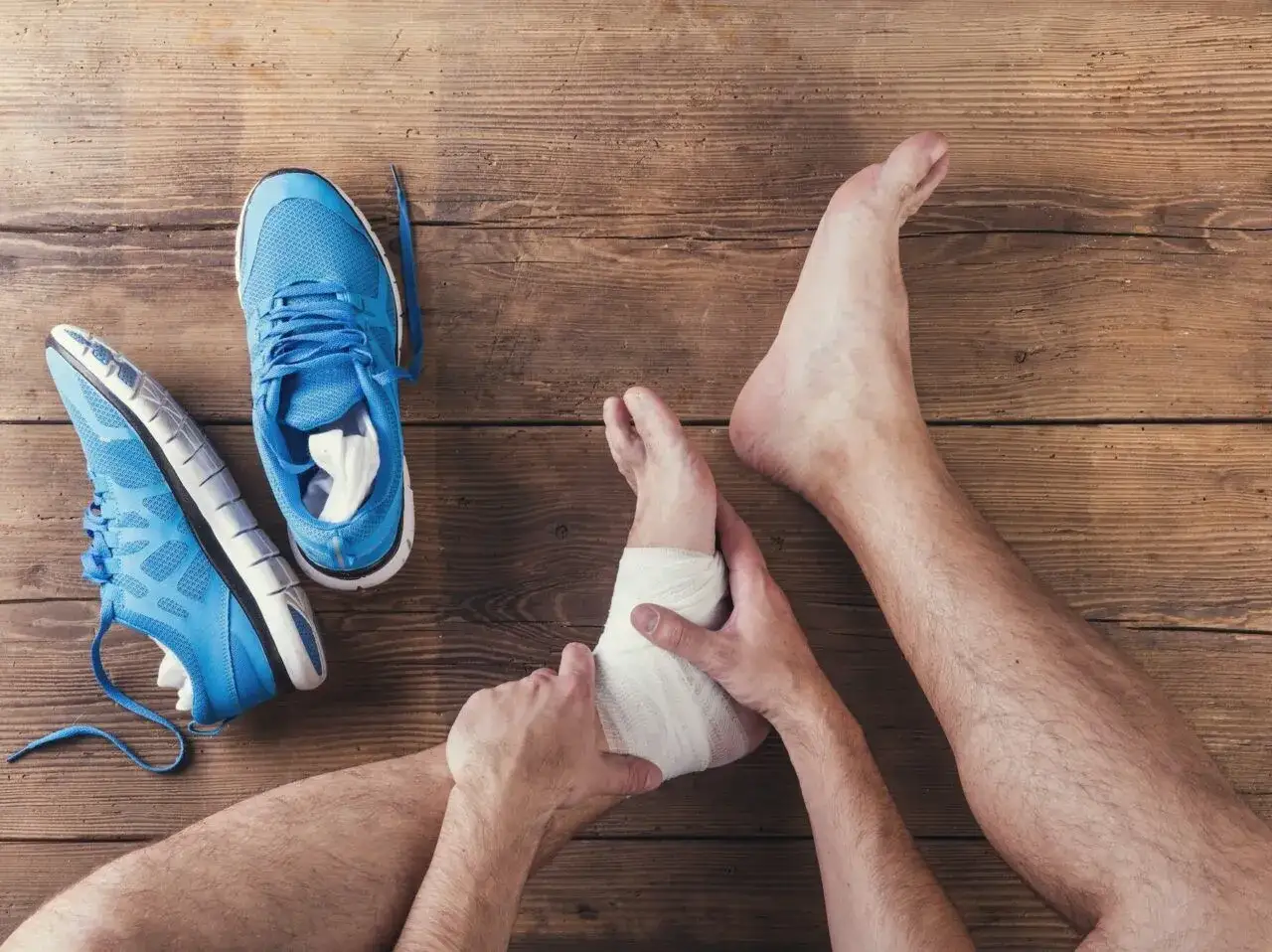 Opatrywanie skręconej kostki bandażem, obok leżą niebieskie buty sportowe. To pierwszy krok, jak pozbyć się opuchlizny po skręceniu kostki.