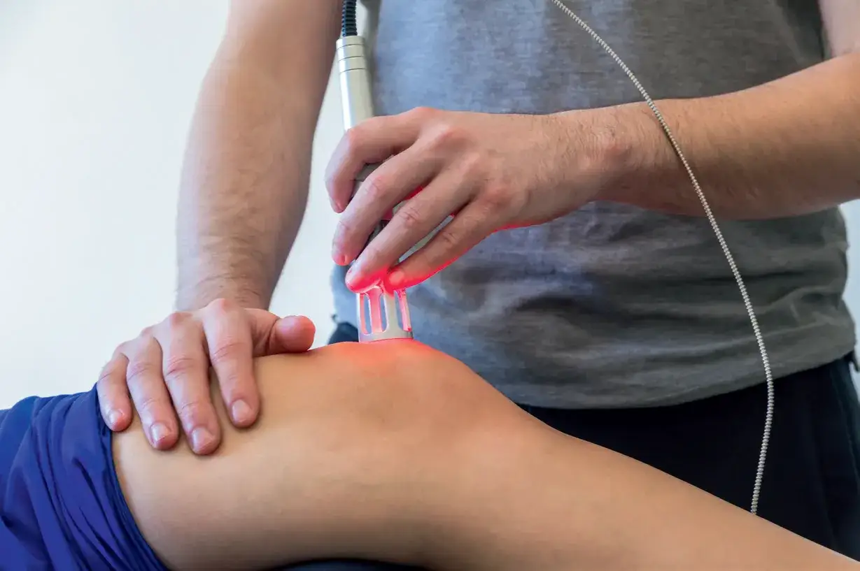 Rehabilitacja laserem na czym polega? Terapeuta naświetla kolano pacjenta czerwonym światłem lasera, wspomagając regenerację.