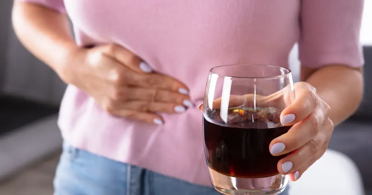 Kobieta trzyma szklankę z ciemnym napojem, odczuwając ból brzucha po alkoholu. Co robić w takiej sytuacji?